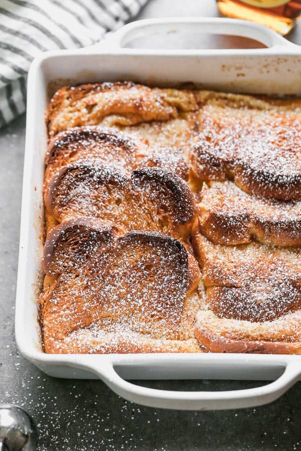 Baked French toast in a white baking pan with powdered sugar over top. 