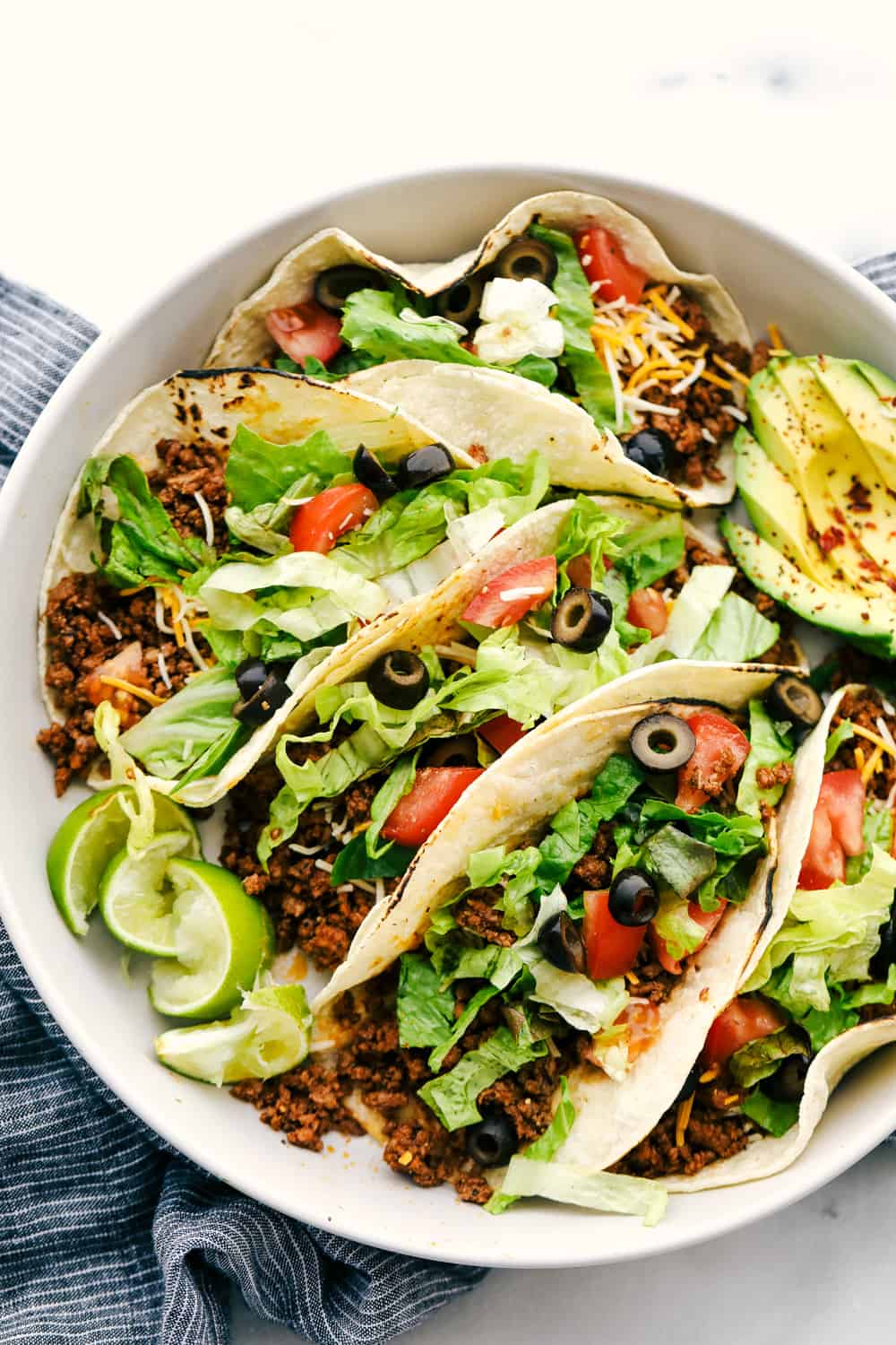 A plate of soft tacos grilled with meat and topped with shredded cheese, olives, lettuce, tomatoes and sliced avocados. 