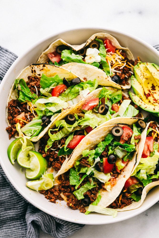 Five beef tacos on a plate loaded with shredded lettuce, sliced black olives, shredded cheese, tomato slices with lime slices and an avocado sliced on the side. 