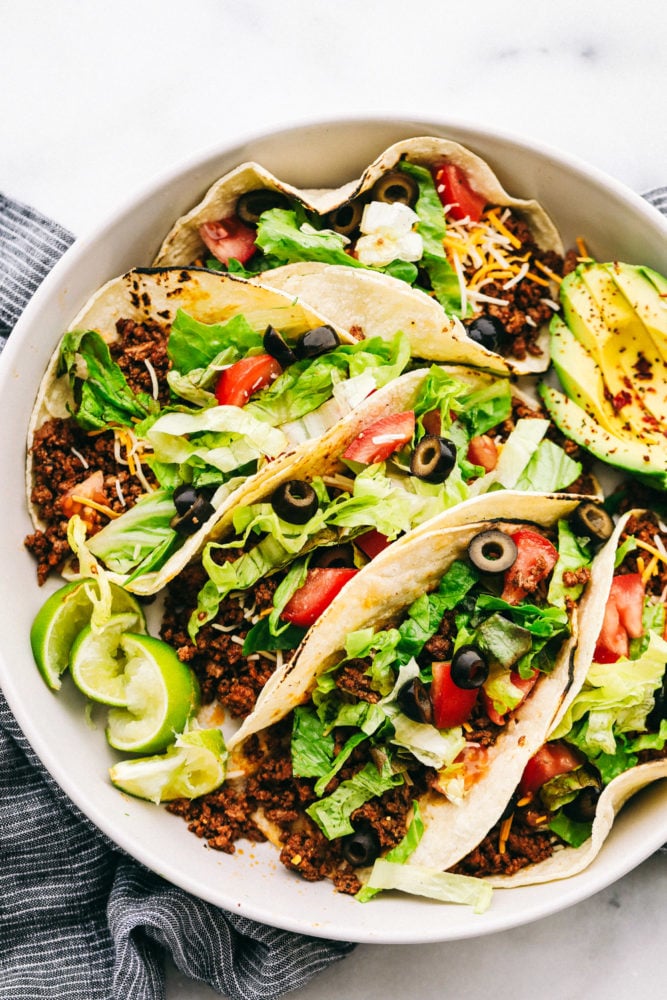 Cinco tacos de carne en un plato cargado de lechuga rallada, rodajas de aceitunas negras, queso rallado, rodajas de tomate con rodajas de lima y un aguacate en rodajas a un lado. 