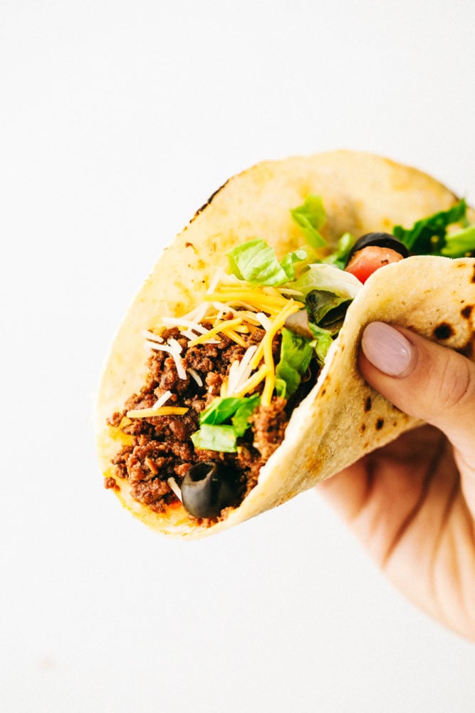 A beef taco with olives, shredded lettuce and shredded cheese being held up with the recipe critic hand. 