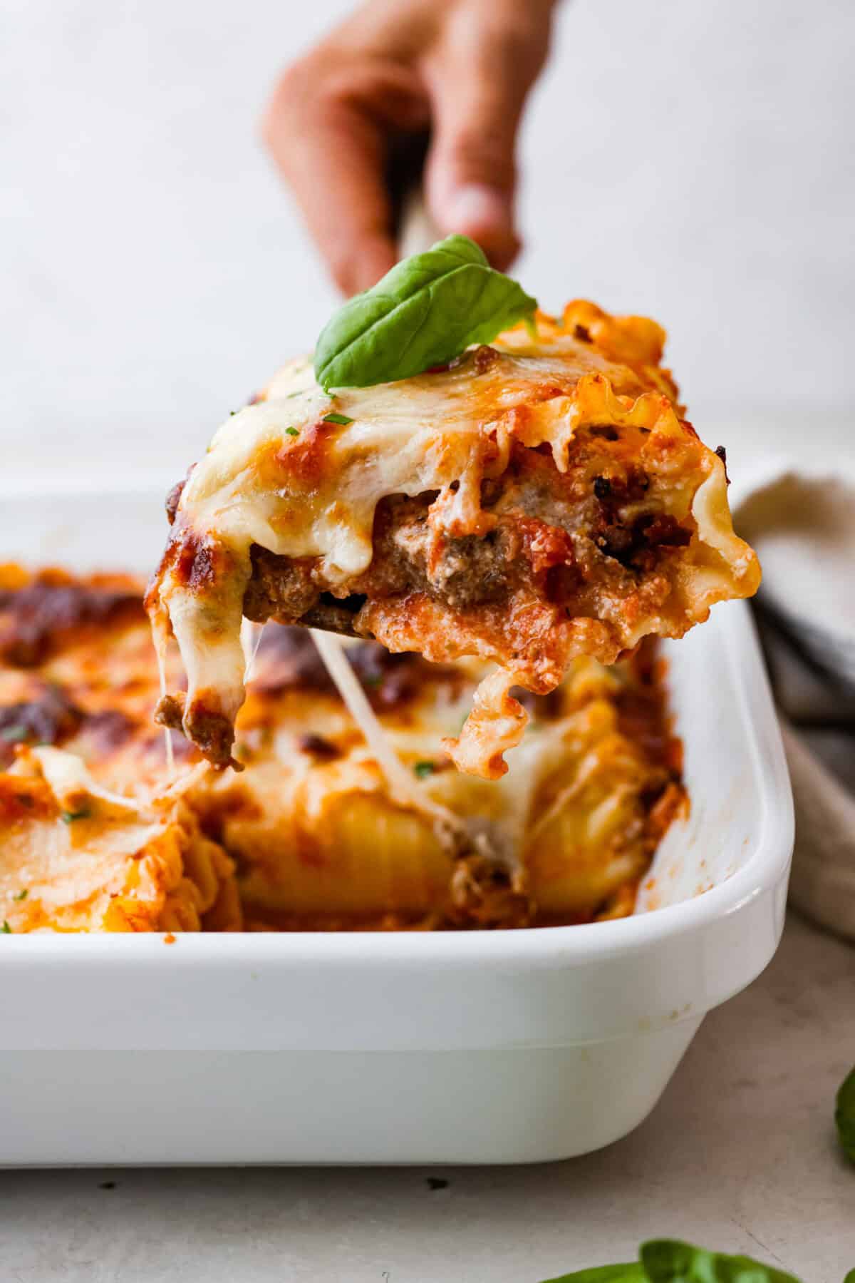 Side shot of someone lifting a lasagna roll up with a basil leaf on top, out of the baking dish. 