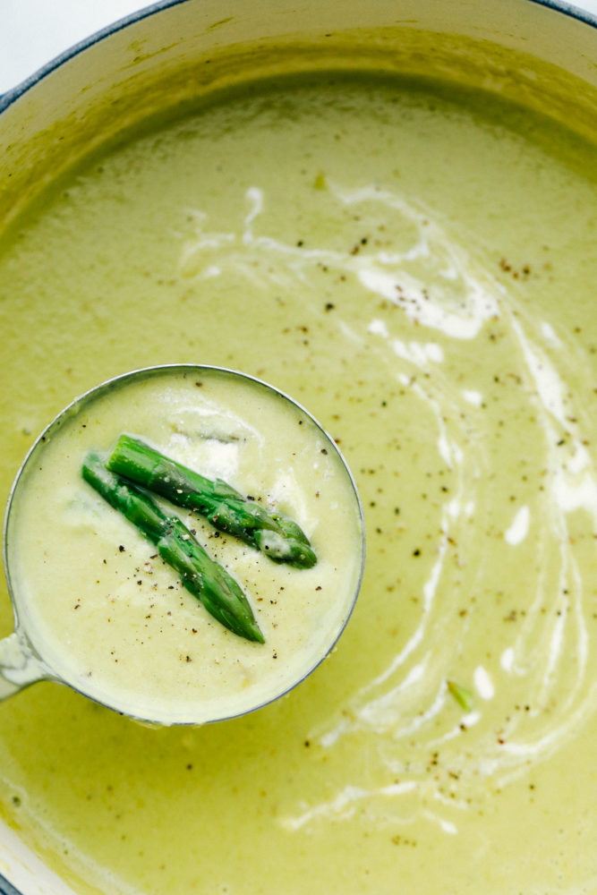 Asparagus soup in a pot with a ladle full of asparagus soup and two asparagus tips. 