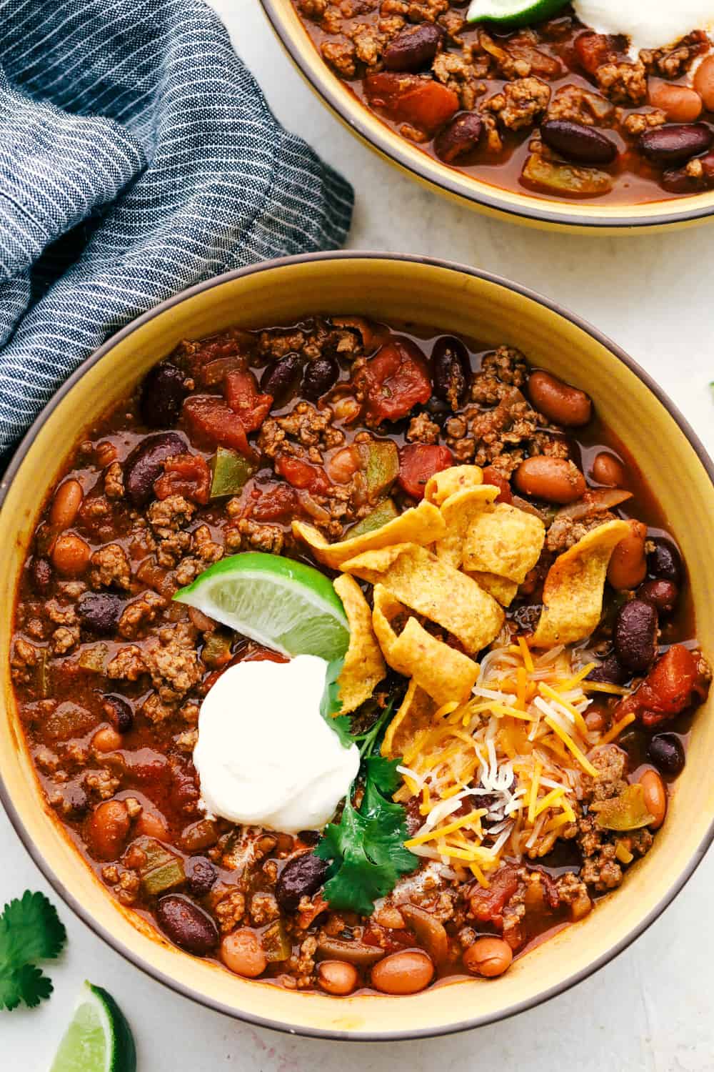 Chili in a bowl topped with lime wedge, sour cream, shredded cheese and Frito chips. 