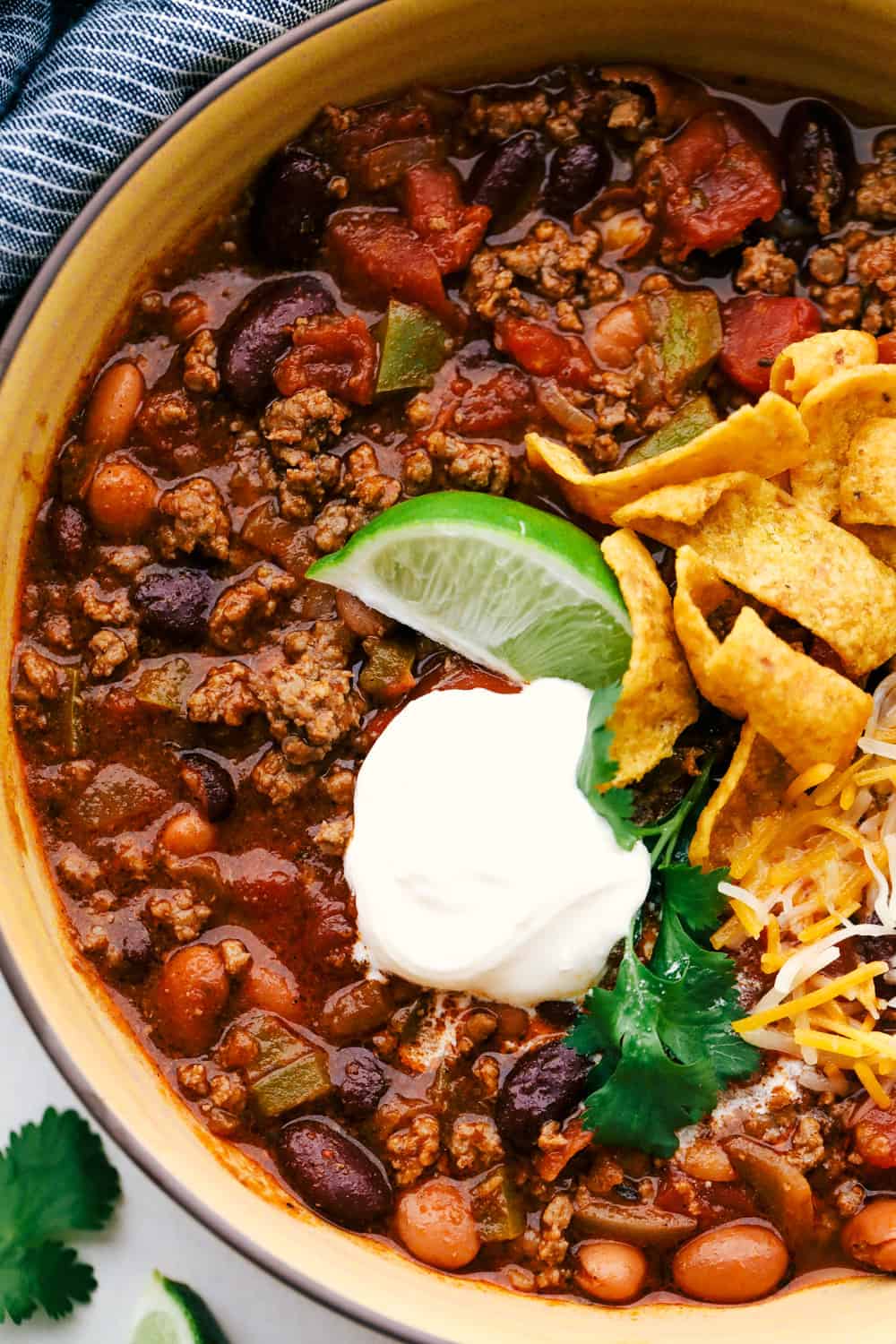 Chili in a bowl with lime wedge, sour cream, Frito chips and shredded cheese. 