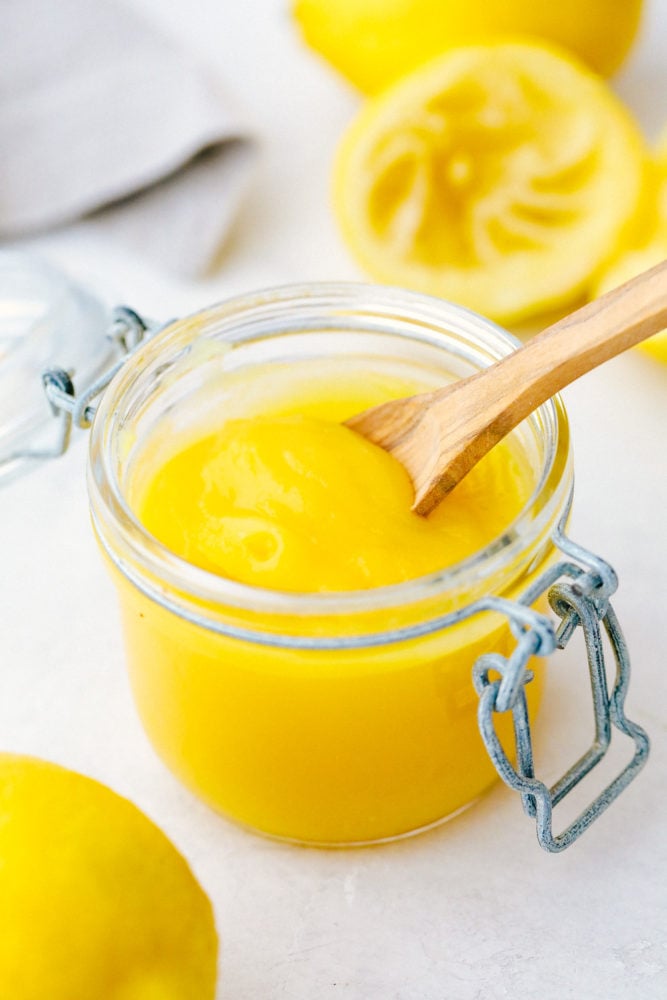 Lemon curd in an open jar with a small wooden spoon and lemons laying next to the jar as decoration. 