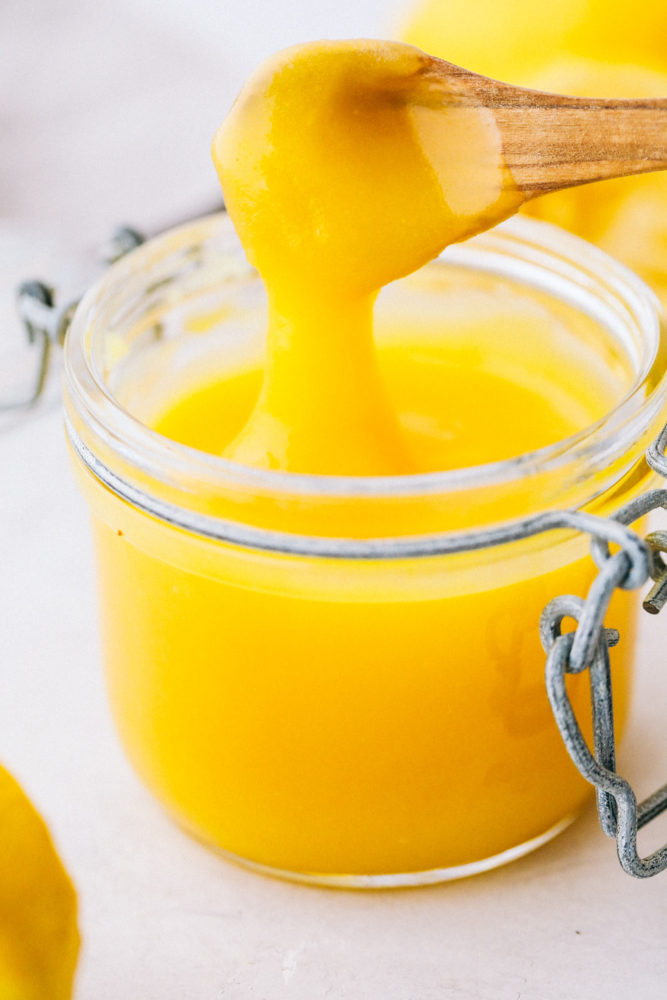 Lemon curd in a small jar with a small wooden spoon with lemon curd dripping off the spoon. 