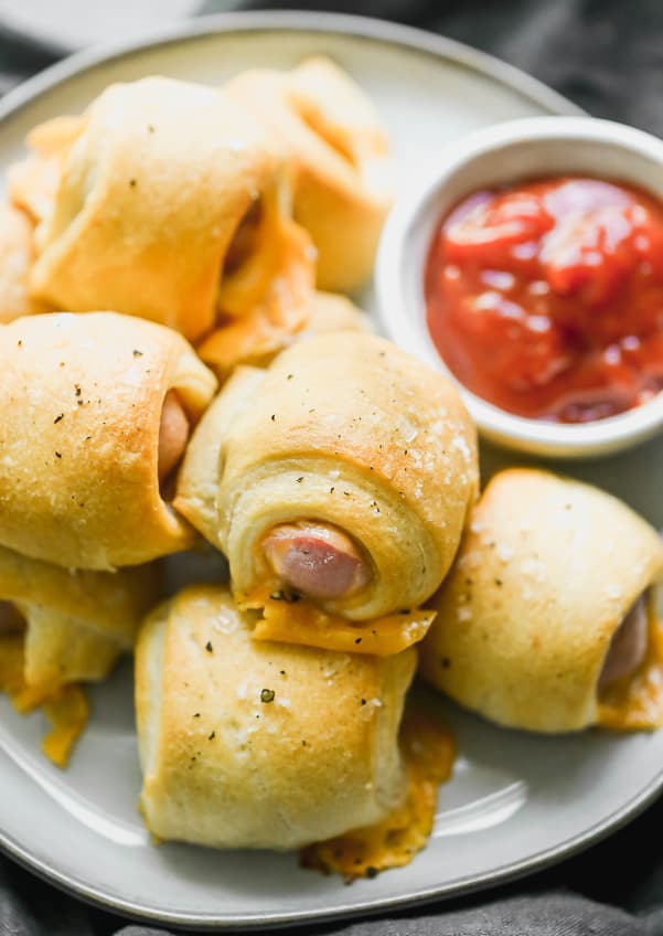 Pigs in a blanket stacked up on a white plate with marinara sauce on the side. 
