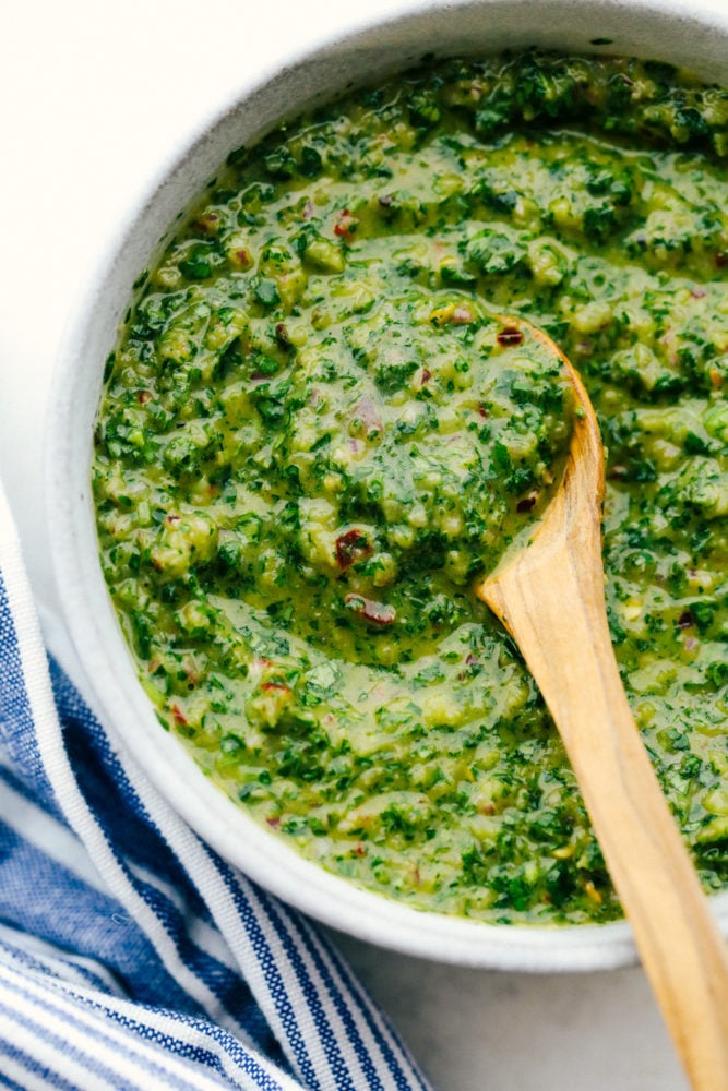 Chimichurri sauce in a white bowl with a wooden spoon stirring it. 