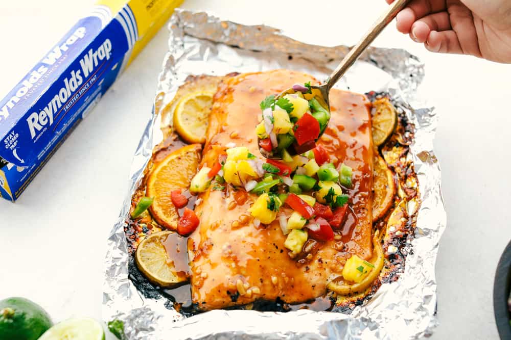 Grilled citrus salmon on Reynolds wrap aluminum foil being garnished with pineapple salsa using a silver spoon to add it to the salmon.
