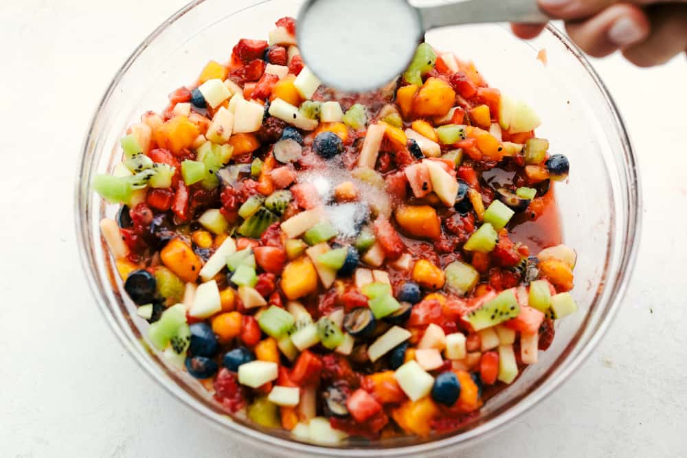 Fruit salsa cut up in a glass bowl with juice being poured over top in a measuring cup. 