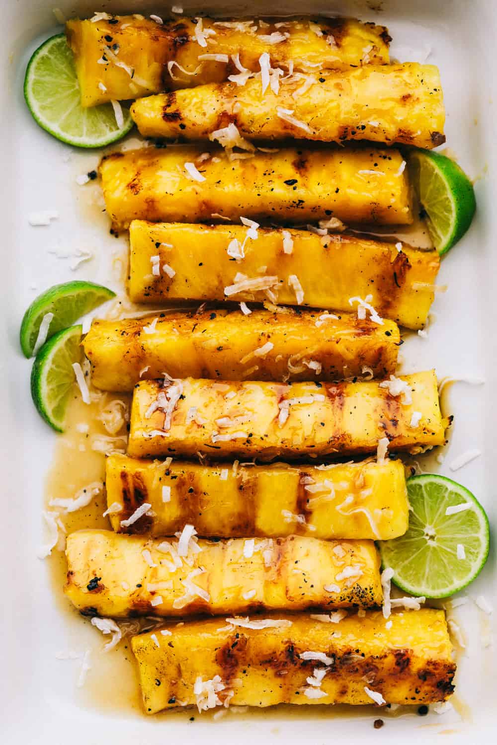 Grilled pineapple in a pan with coconut flakes and lime wedges as garnish. 