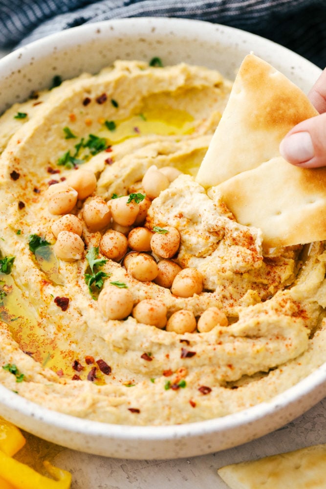 Hummus in a white bowl with a pita chip being dipped in it and chickpeas over top as a garnish. 