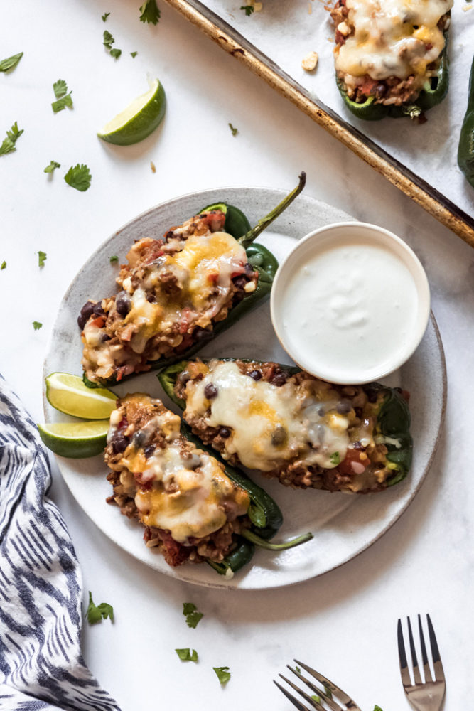 Three stuffed poblano peppers on a plate with a bowl of crema and lime wedges.