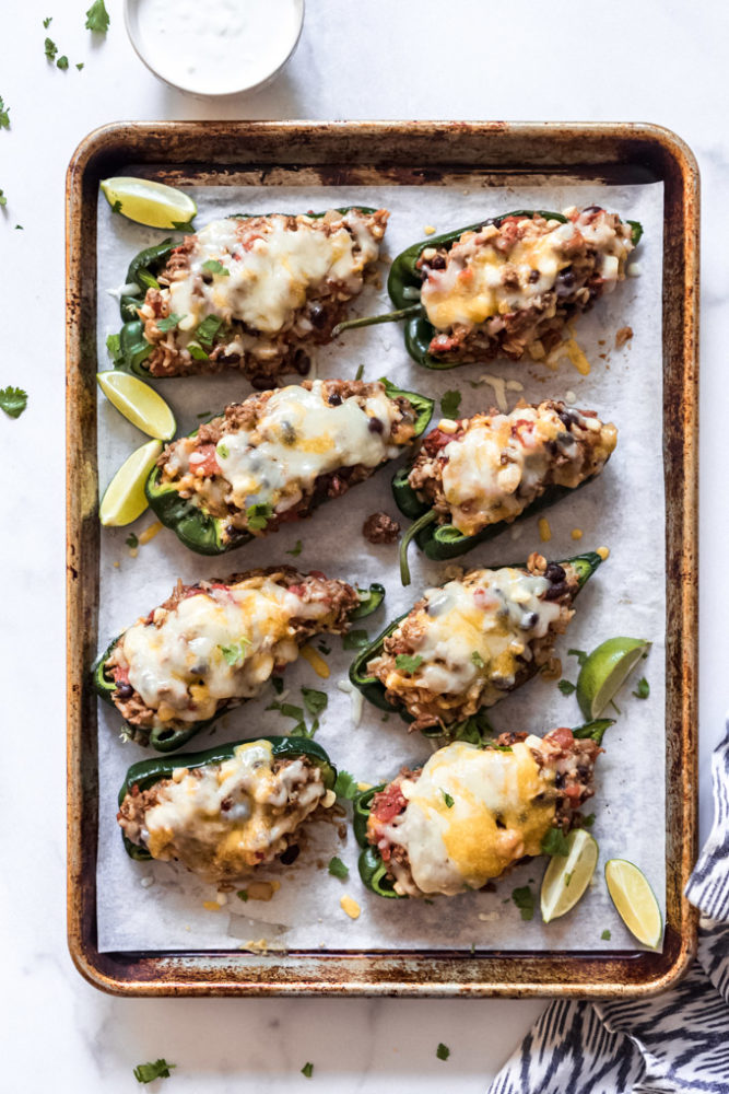Eight stuffed poblano peppers topped with melted cheese on a baking sheet with lime wedges.