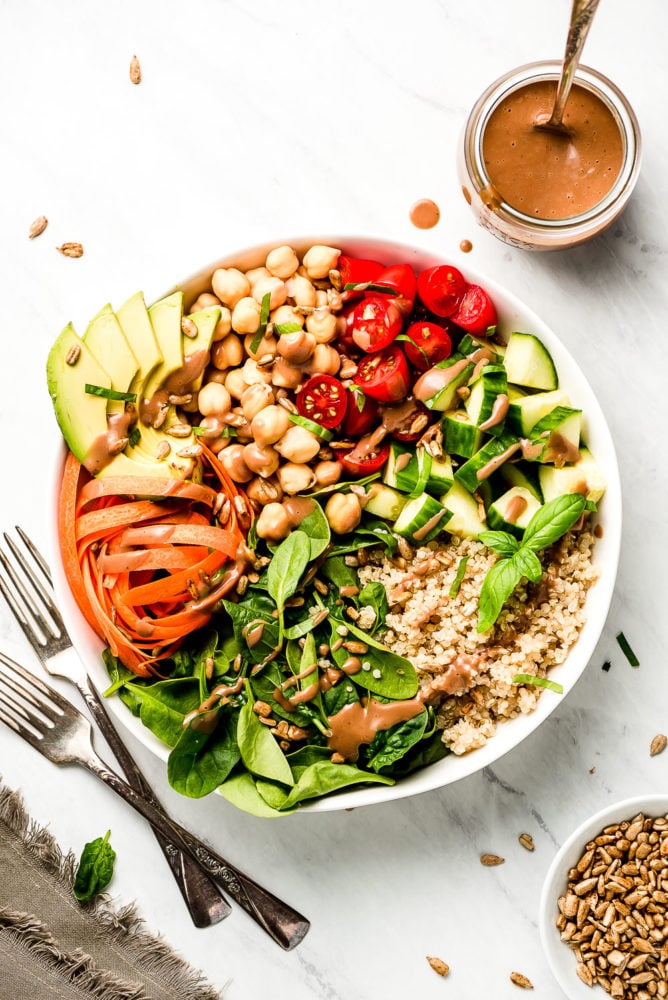 A raw vegetable Vegan Buddha Bowl made up of with spinach, carrots, avocado, chickpeas, grape tomatoes, cucumbers, quinoa, basil, sesame seeds, and a creamy balsamic dressing.