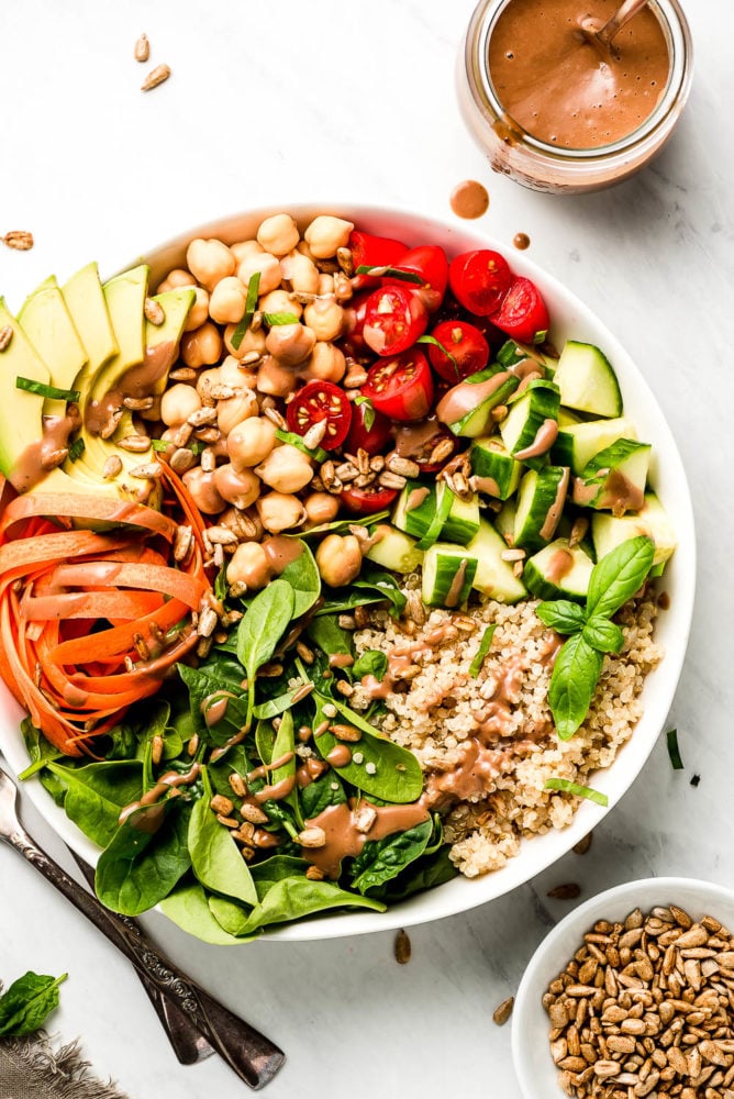 A large Buddha Bowl with spinach, quinoa, carrots, cucumbers, tomatoes, chickpeas, and avocado with a jar or dark creamy dressing to the side.