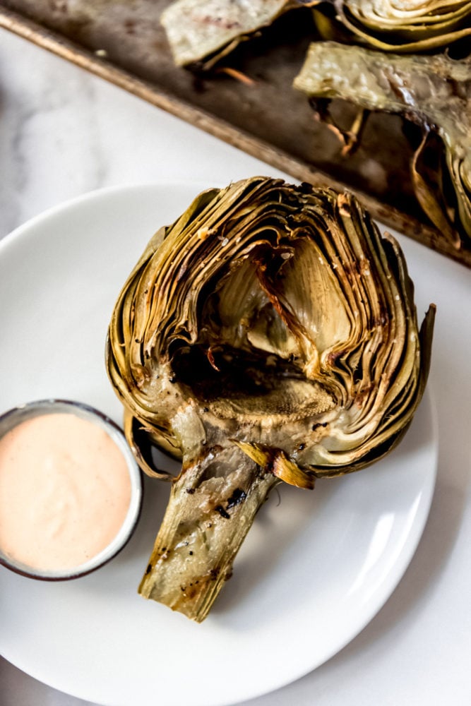 A grilled artichoke half on a white plate with dipping sauce.