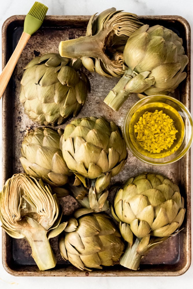 Boiled artichokes on a baking sheet with olive oil and minced garlic for brushing.