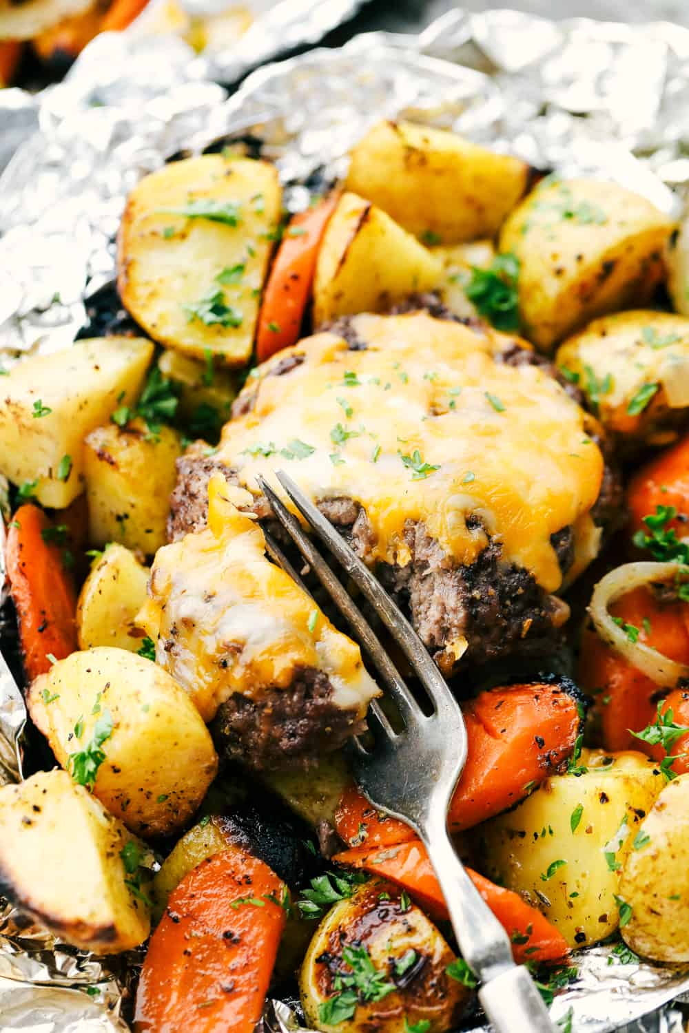 Hobo dinner with foil hamburger being cut with a fork over top of vegetables. 