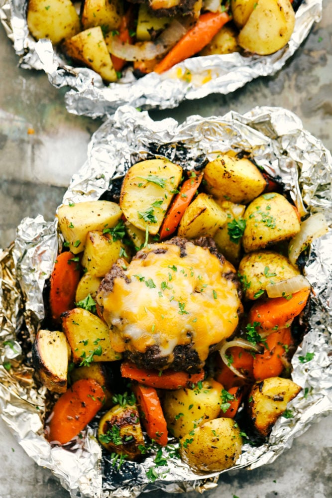 Hobo dinner foil packets opened with vegetables and hamburger patty on top with a slice of cheese. 