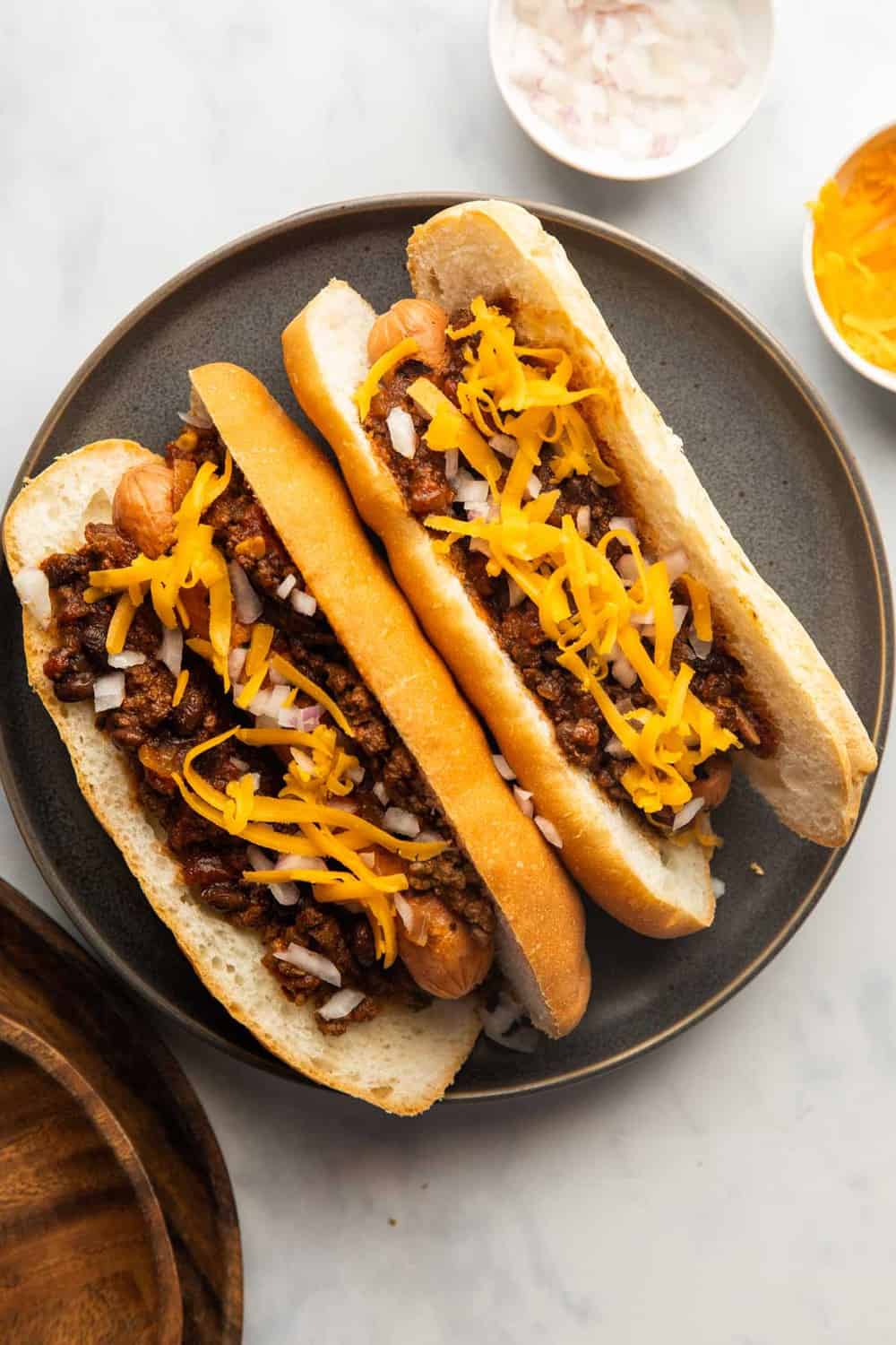 Chili Dogs served on a plate with shredded cheddar and onions on the side