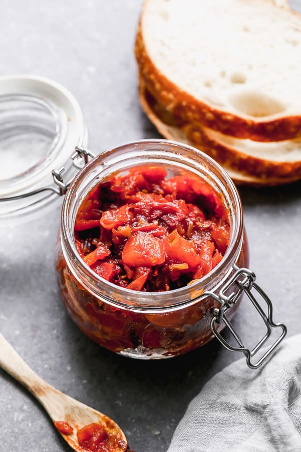 Mermelada de tomate en un frasco transparente con una cuchara de madera y tres rebanadas de pan que rodean el frasco. 