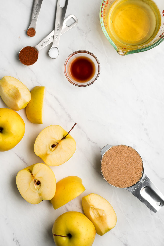 Apples cut in half and quarters, teaspoons of spices, a small bowl of vanilla extract, a liquid measuring cup of apple cider, and a measuring cup of brown sugar.