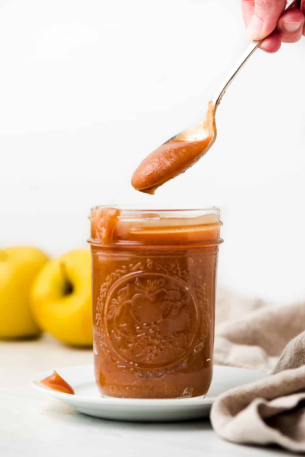 Lifting a spoonful of Apple Butter out of a jar on a plate with apples in the background.