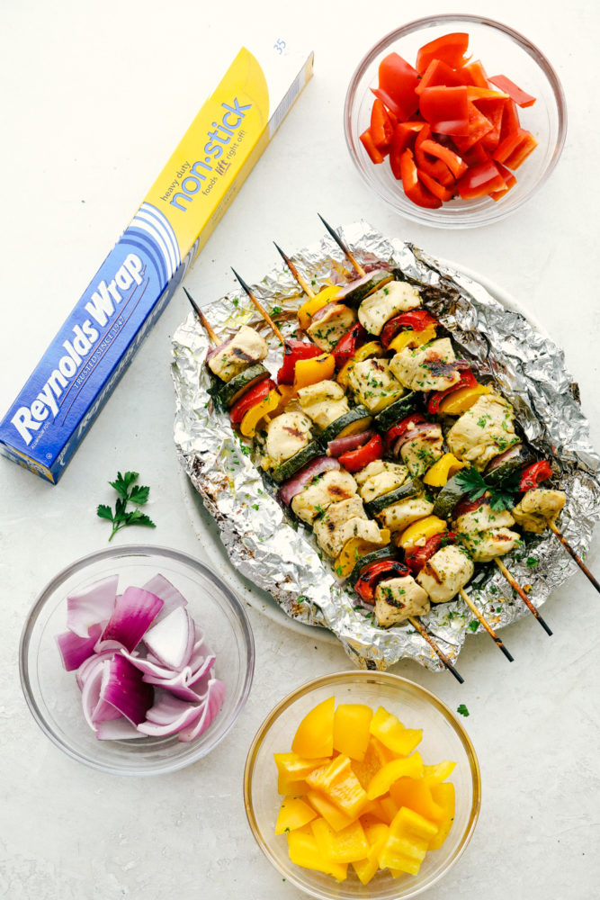 Chicken skewers laying on Reynolds wrap with bell peppers and onions in separate glass bowls. 