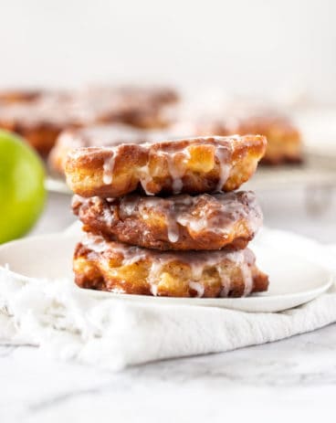 A stack of 3 apple fritters on a white plate with more in the background