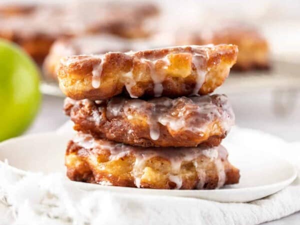 A stack of 3 apple fritters on a white plate with more in the background