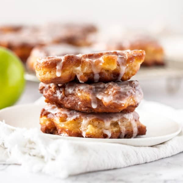 A stack of 3 apple fritters on a white plate with more in the background