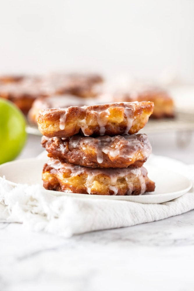 Una pila de 3 buñuelos de manzana en una placa blanca con más en el fondo