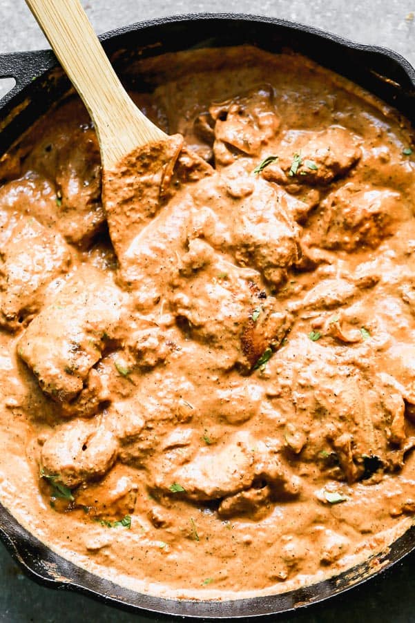 Butter chicken in a black skillet being stirred by a wooden spoon. 