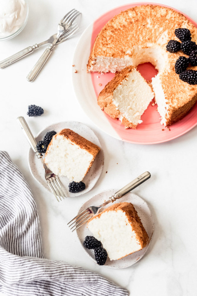 Slices of angel food cake on dessert plates with blackberries and forks next to the rest of the cake on a pink plate.