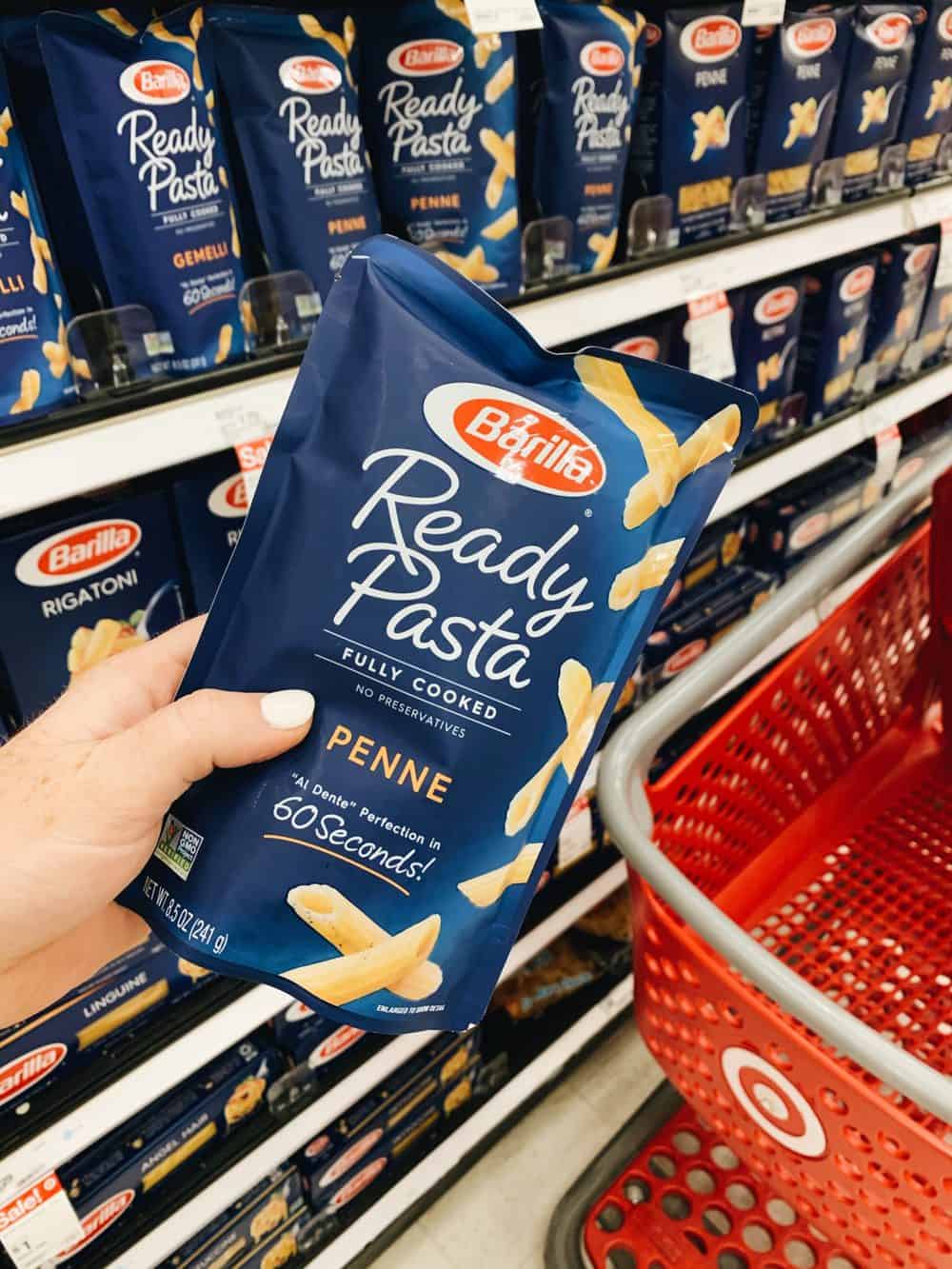 Ready pasta being held up in front of other ready pasta packages and a target shopping cart. 