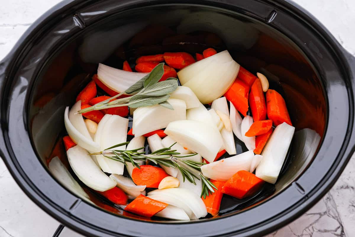 Slow Cooker Turkey Breast is the perfect way to get tender, juicy turkey with a fraction of the prep but all the flavor. Let the slow cooker do ALL the work! Overhead shot of onion, carrots, garlic and sprigs of herbs in the bottom of the crockpot.