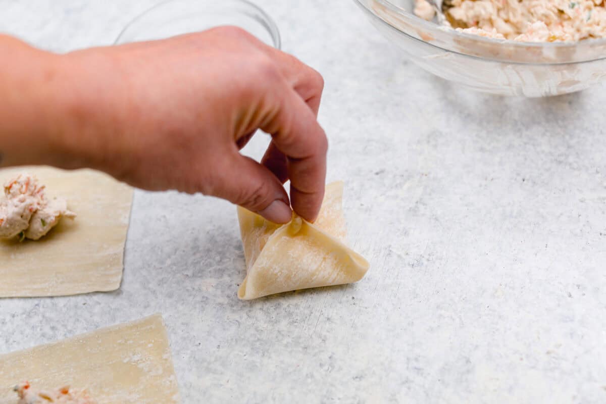 Angle shot of someone pinching all four corners of the wonton paper together with filling inside. 