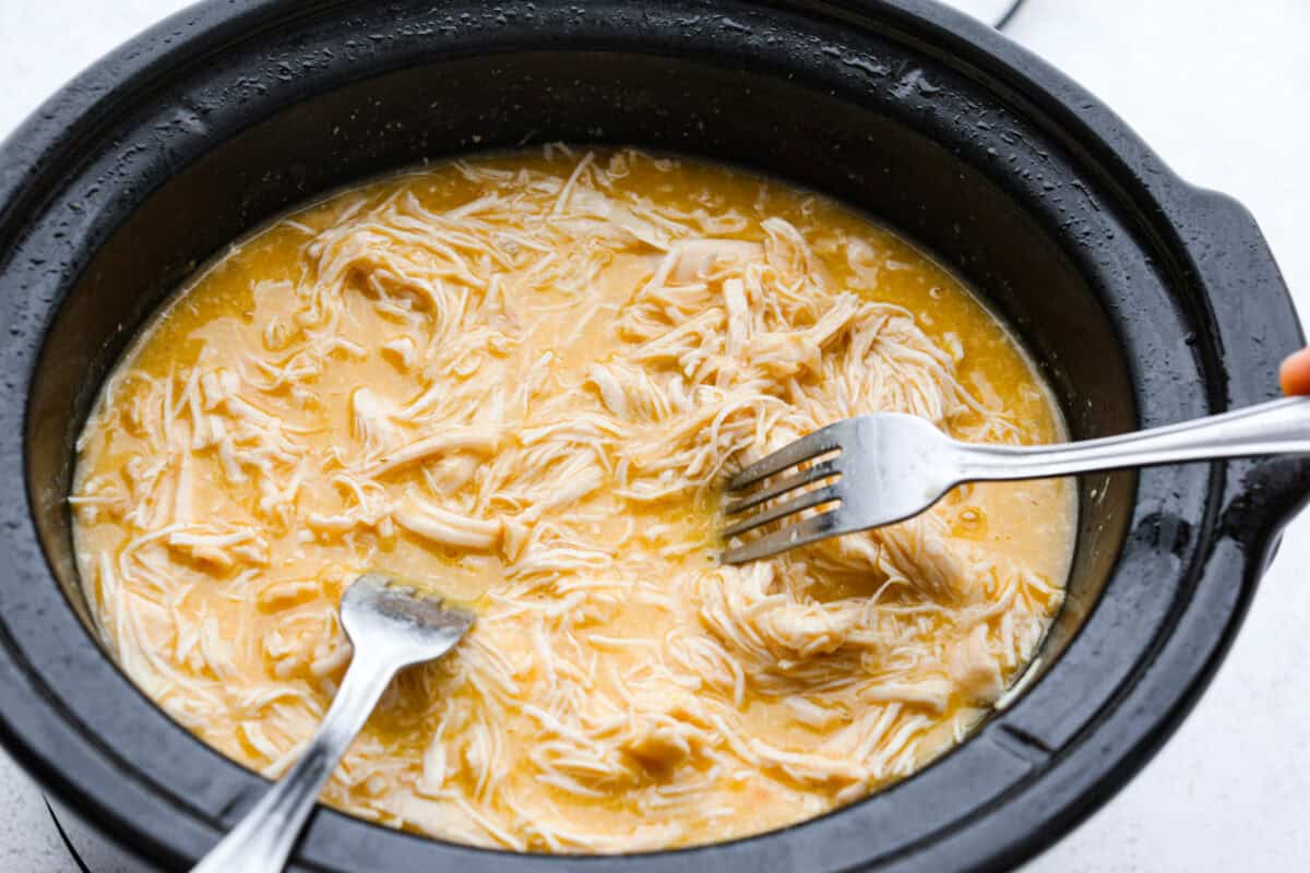 Overhead shot of someone shredding chicken with two forks in sauce in the crockpot. 