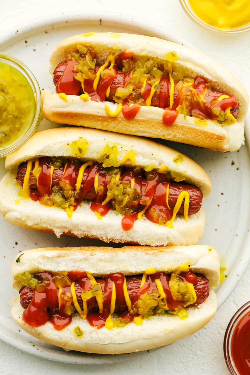 Air fryer hot dogs in a bun with mustard, ketchup and relish on a plate. 