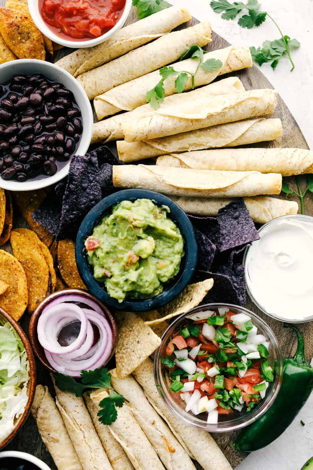 Taquitos on a board with bowls of dips, vegetables and chips all around. 