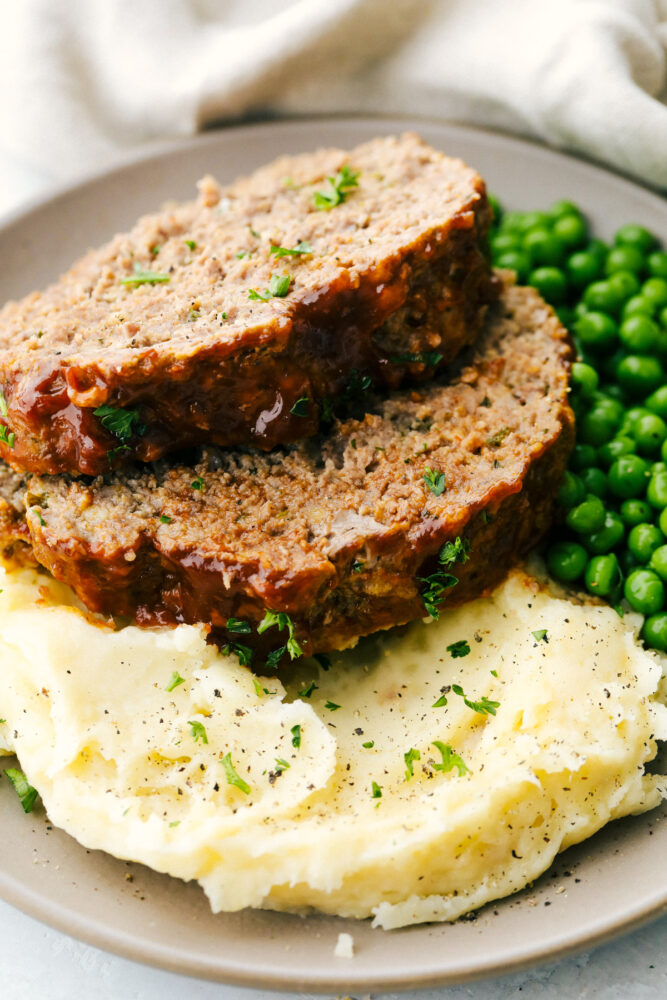 Sliced meatloaf on a plate with a side of peas and mashed potatoes. 