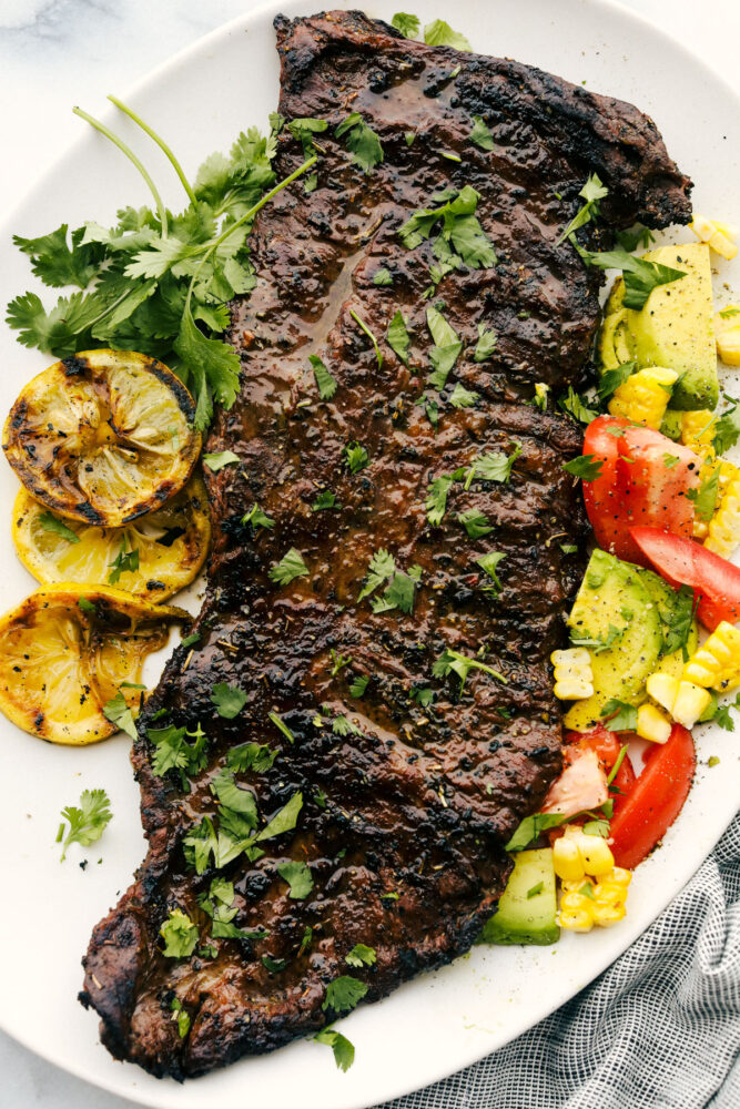 The grilled skirt steak whole on a plate with tomatoes, corn and tomatoes. 