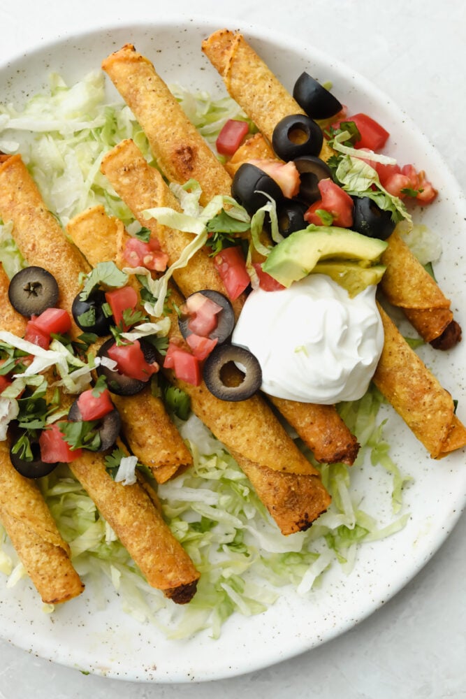 Air fryer taquitos on a white plate with sour cream, olives, tomatoes and lettuce.