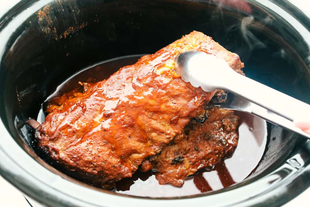 Someone taking cooked ribs out of the slow cooker with tongs.