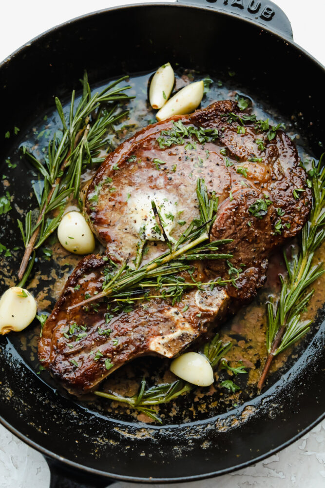 Cooked ribeye steak in a skillet, garnished with garlic, rosemary, and thyme.