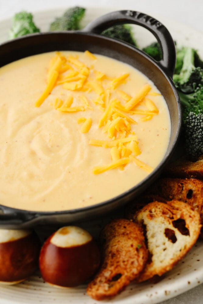 Beer cheese dip on a platter with broccoli, pretzel rolls and a crostini. 