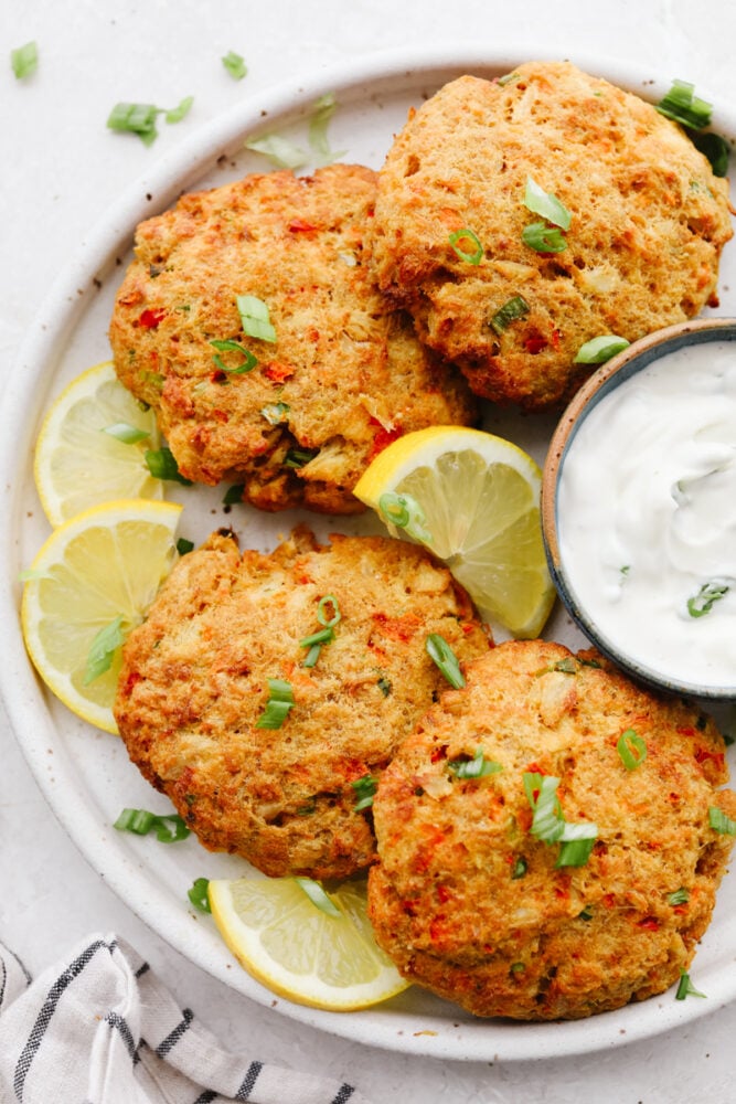 4 air fryer crab cakes on a white plate with lemon and green onion garnish.