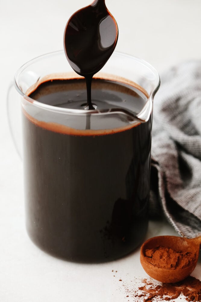 A spoon being pulled out of a pitcher filled with chocolate sauce. 